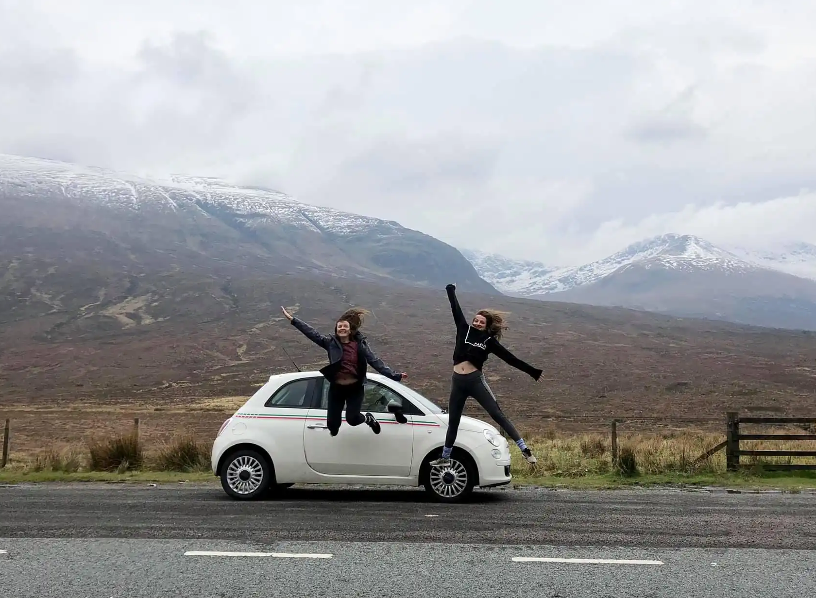 Girls jumping with a fiat 500 parked and the Scottish Highlands in the background.