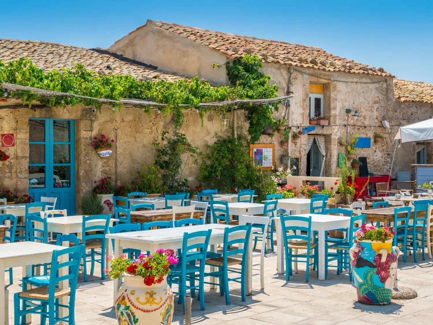 Restaurant with colourful outdoor seating and rustic house in the background in Marzamemi, Sicily.